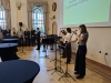 Trzy artystki występują na scenie w jasnej, klasycznej sali; jedna śpiewa do mikrofonu, druga gra na skrzypcach, a trzecia stoi przy pulpicie nutowym. W tle, pod dużym ekranem z noworocznymi życzeniami, stoi grupa osób w strojach wizytowych, czarny fortepian oraz wysokie stoliki bankietowe. Całość odbywa się w eleganckim wnętrzu z wysokimi oknami i ozdobnym parkietem, co nadaje wydarzeniu uroczysty charakter.