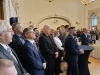 Grupa mężczyzn i kobiet w strojach wizytowych stoi w rzędzie, z uwagą słuchając trwającego wystąpienia. Na pierwszym planie widoczne są profile twarzy uczestników, a w tle rozciąga się przestronna sala z bufetem i kolejnymi grupami gości. Oświetlenie z żyrandoli i jasne ściany podkreślają powagę oraz elegancję uroczystości.