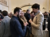Uczestnicy spotkania rozmawiają w grupach, trzymając w dłoniach kieliszki z napojami w wypełnionej ludźmi sali bankietowej. Na pierwszym planie widać dwoje mężczyzn i kobietę zaangażowanych w dyskusję, a w tle innych gości oraz fragment bufetu z filiżankami. Ciepłe oświetlenie i klasyczne detale architektoniczne podkreślają swobodną, a zarazem kulturalną atmosferę wydarzenia.