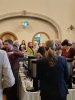 Goście gromadzą się wokół długiego stołu bufetowego, na którym ustawione są termosy z kawą i herbatą oraz dzbanki z dodatkami. Centralną część ujęcia zajmuje tył sylwetki kobiety w ciemnej marynarce, która patrzy w stronę tłumu rozmawiającego w głębi sali pod ozdobnym łukiem. Wnętrze oświetla rzutnik zamontowany pod sufitem, a w tle widoczne są lustra i detale architektoniczne eleganckiej auli.