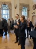 Kobieta w okularach przemawia do mikrofonu, trzymając w drugiej ręce kieliszek, podczas gdy obok niej stoi uśmiechnięty mężczyzna w garniturze. W tle widać pozostałych gości oraz stylowe wnętrze auli z wysokimi oknami i drewnianymi drzwiami. Atmosfera spotkania jest oficjalna, ale jednocześnie serdeczna i radosna.