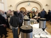 Licznie zgromadzeni goście korzystają z poczęstunku przygotowanego na długim stole, nakładając ciasta na białe talerzyki. Na pierwszym planie znajdują się duże termosy gastronomiczne oraz starannie ułożone rzędy czystych filiżanek. W głębi sali uczestnicy prowadzą swobodne rozmowy w jasnym, przestronnym wnętrzu zwieńczonym dekoracyjnym łukiem.