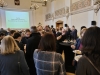 Uczestnicy spotkania, zwróceni tyłem i bokiem, wypełniają przestronną salę, jedząc posiłki i rozmawiając przy okrągłych stolikach. Nad tłumem dominuje duży ekran z wyświetlonymi zielonymi literami życzeniami noworocznymi oraz polskie godło na ścianie powyżej. Wysokie ściany zdobią klasyczne freski oraz ozdobne żyrandole zwisające z sufitu.