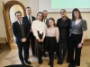 Grupa siedmiu osób w strojach wizytowych pozuje wspólnie do zdjęcia na tle jasnej ściany i fragmentu ekranu projekcyjnego. Wśród uśmiechniętych uczestników znajdują się zarówno kobiety, jak i mężczyźni, w tym artystki występujące wcześniej na scenie. Elegancki ubiór i geometryczny wzór parkietu pod ich stopami podkreślają uroczysty charakter zakończenia części oficjalnej.