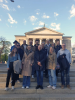 Grupa kilkunastu uśmiechniętych osób pozuje do wspólnej fotografii na szerokich, kamiennych schodach. W tle widoczny jest monumentalny budynek z klasycznymi kolumnami i trójkątnym frontonem, zwieńczony rzeźbą na dachu. Uczestnicy są ubrani w codzienne, wiosenne stroje, takie jak kurtki, płaszcze i swetry, a nad nimi rozciąga się jasne, błękitne niebo z delikatnymi chmurami. Całość kompozycji ma charakter pamiątkowego zdjęcia wykonanego przed zabytkowym obiektem w świetle dziennym.