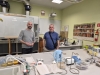 Dwóch mężczyzn stoi w laboratorium naukowym wypełnionym specjalistycznym sprzętem i pomocami dydaktycznymi. Pierwszy plan zajmuje stół laboratoryjny, na którym znajdują się pojemniki z próbkami, naczynia szklane z kolorowymi płynami oraz błękitny czajnik. W tle widoczna jest biała tablica, godło państwowe, mapy geologiczne oraz system wentylacyjny pod sufitem. Panowie patrzą w stronę obiektywu, jeden z nich ma na ramieniu przewieszoną torbę, a drugi trzyma w dłoniach mały przedmiot.