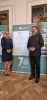Kobieta w ciemnej sukience oraz mężczyzna w brązowym mundurze leśnika stoją obok informacyjnego roll-upu. Baner upamiętnia 70 lat współpracy Biura Urządzania Lasu i Geodezji Leśnej w Brzegu z Wydziałem Leśnym i Technologii Drewna UPP. Para uśmiecha się, splatając dłonie przed sobą na tle klasycznego wnętrza z drewnianym parkietem i obrazami na ścianach. Postacie ustawione są symetrycznie po obu stronach zielono-białej planszy z logotypem BULiGL.