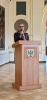 Kobieta w okularach i ciemnej sukience przemawia do mikrofonu, stojąc za drewnianą mównicą. Na froncie pulpitu widnieje herb powiatu brzeskiego z napisem „Powiat Brzeski” oraz symbolem czarnego orła. Wystąpienie odbywa się w jasnej, zabytkowej sali z ozdobnymi detalami na ścianach i klasycznym obrazem w tle. Postać uchwycona jest w zbliżeniu podczas oficjalnej części wydarzenia.