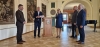 Grupa pięciu osób uczestniczy w oficjalnym spotkaniu w eleganckiej sali bankietowej zdobionej klasycznymi obrazami. W centralnym punkcie kobieta przemawia do mikrofonu, stojąc obok drewnianej mównicy z herbem przedstawiającym czarnego orła. Pozostali uczestnicy, ubrani w garnitury, obserwują wystąpienie, a jeden z mężczyzn trzyma w dłoniach płaskie, ozdobne pudełko. W tle widoczne są wysokie arkady prowadzące do kolejnych pomieszczeń oraz fragment czarnego fortepianu.