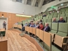 Kobieta w szarym kostiumie stoi przodem do publiczności i mówi do mikrofonu na dole dużej sali wykładowej. Słuchacze siedzą na zielonych fotelach, które są ustawione rzędami i pną się wysoko w górę. Pomieszczenie ma drewnianą podłogę oraz ściany, a wysoki sufit i okna znajdują się po lewej stronie. Wszyscy zebrani uważnie patrzą w stronę prowadzącej spotkanie.